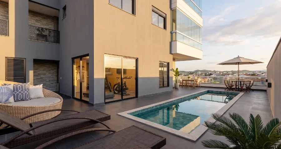 Deck da piscina do Residencial AltaVille ao pôr do sol, com espreguiçadeiras, ombrelone e a academia envidraçada ao lado