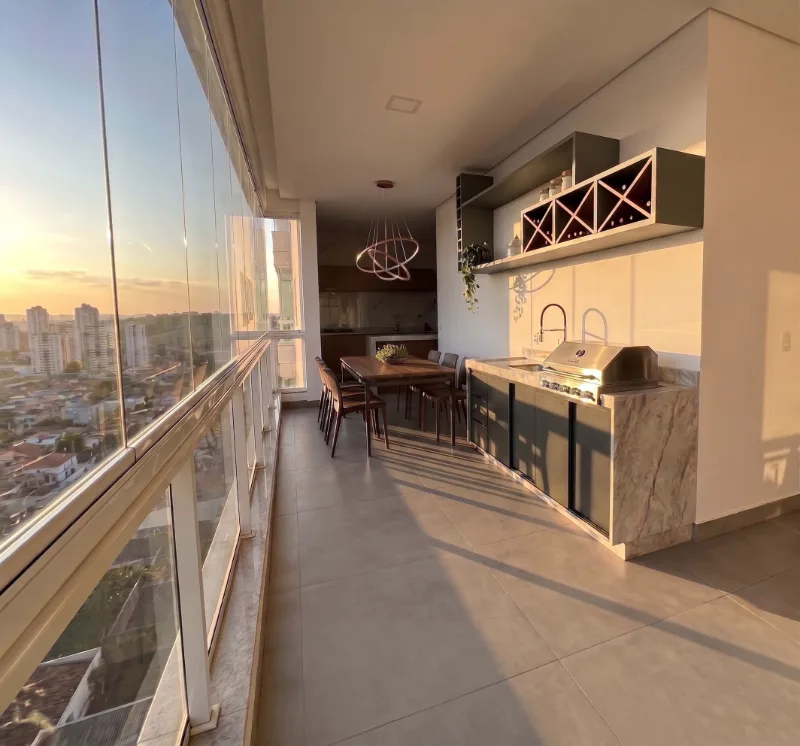 Varanda gourmet do apartamento, integrada à área social, com amplas esquadrias de vidro do chão ao teto e vista para a cidade ao pôr do sol